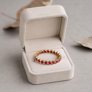 May include: A gold eternity ring featuring a continuous band of small, round, vibrant pinkish-red gemstones set in individual metal bezels. The ring is displayed inside an open, light beige, velvet-lined jewelry box with a hinged lid.