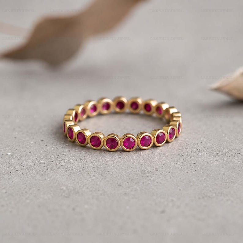 May include: A gold eternity band featuring a continuous row of round, bezel-set vibrant pink gemstones. The ring has a polished metallic finish and a uniform circular design, showcasing a series of identical stones set securely within the gold metal.