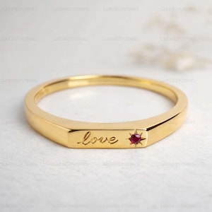 Peut inclure: Une bague en or avec le mot "love" gravé sur le bandeau. Une petite pierre précieuse rouge est sertie dans un motif d'éclat d'étoile. La bague a une finition polie et un design simple et élégant.