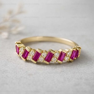 Peut inclure: Une bague en or ornée de pierres magenta rectangulaires alternées et de petites pierres claires rondes. La bague a un anneau délicat et un motif en zigzag de pierres précieuses. La bague est posée sur une surface gris clair.