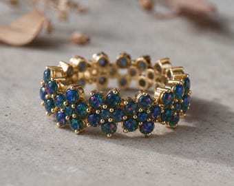 May include: A gold ring featuring a floral design with multiple dark blue and green gemstones. The ring is set on a gray surface, showcasing the intricate details of the jewelry.