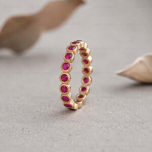 May include: A gold eternity band featuring a continuous row of round, vibrant pink rubies set in individual circular bezels. The ring displays a uniform design with each gemstone securely held in place by the polished gold metal setting.