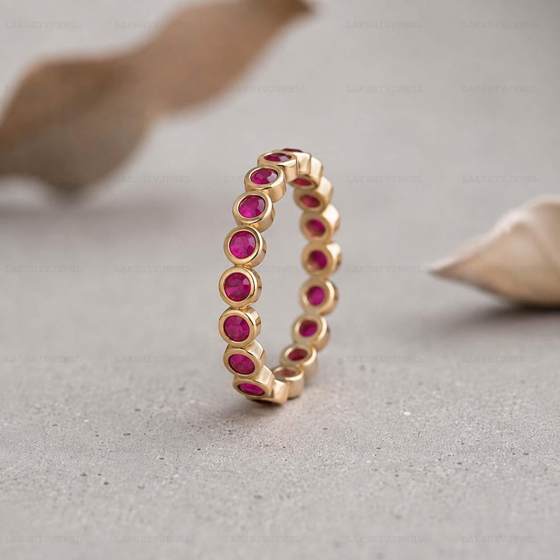 May include: A gold eternity band featuring a continuous row of round, vibrant pink rubies set in individual circular bezels. The ring displays a uniform design with each gemstone securely held in place by the polished gold metal setting.