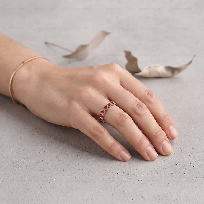 May include: A gold band ring featuring a series of small, round, bezel-set red rubies encircling the finger. The ring is worn alongside a thin, plain gold bangle bracelet, both showcasing a polished metallic finish and vibrant red gemstones.