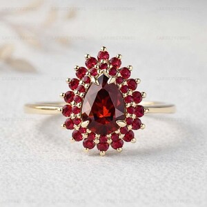 Puede incluir: Un anillo de oro con una gran piedra preciosa roja oscura en forma de pera, rodeada de piedras rojas más pequeñas. El diseño del anillo se asemeja a una explosión floral, con la piedra central como punto focal. La banda es de oro pulido simple.