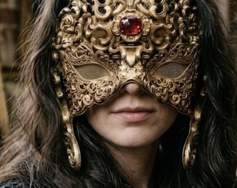 May include: A gold-colored ornate mask featuring intricate scrollwork and filigree patterns. The mask includes a prominent central red gemstone and a smooth gold oval accent above it, covering the upper face with mesh eye openings.