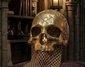 May include: A gold-colored skull sculpture featuring an intricate black symbol on the forehead, dark mesh eye sockets, and a metallic gold chainmail drape hanging from the jaw, resting on a dark velvet cushion with decorative rope trim.