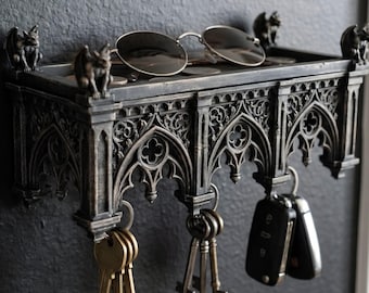 May include: A dark gray wall-mounted shelf featuring intricate gothic architectural arches and small gargoyle figures at the corners. The top surface holds a pair of round sunglasses, while metal hooks underneath display various keys and key fobs.