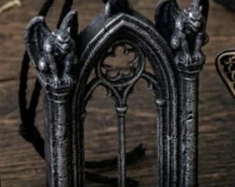 May include: A silver-toned pendant shaped like a miniature arched window frame with a gargoyle perched on each side. The piece features intricate stone-like detailing and is suspended from a thin black cord, creating a detailed architectural accessory.