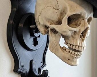May include: A wall-mounted black coat hook featuring a decorative plaque and a human skull wearing a black fedora. The skull is positioned above a curved hook, which holds a piece of black fabric. The entire assembly is mounted on a flat surface.