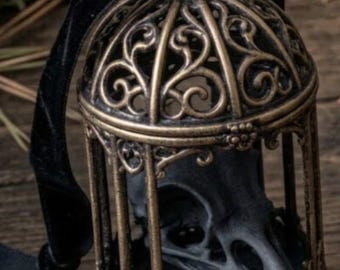May include: A bronze-colored metal birdcage ornament featuring intricate scrollwork and a domed top. Inside the cage sits a matte black bird skull. A black velvet ribbon is attached to the top loop for hanging the decorative object.