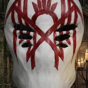 May include: A white full-face mask featuring a hand-painted red symbol across the forehead and eyes. The design includes vertical red brushstrokes extending from the top and sides, with four horizontal eye openings cut into the smooth surface.