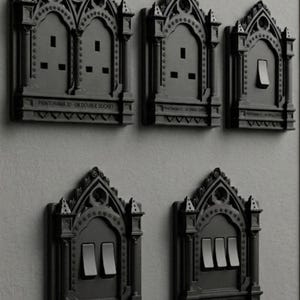 May include: A collection of five matte black electrical wall plates featuring intricate gothic architectural detailing, including pointed arches and columns. The plates include various configurations of power outlets and light switches with text labels.