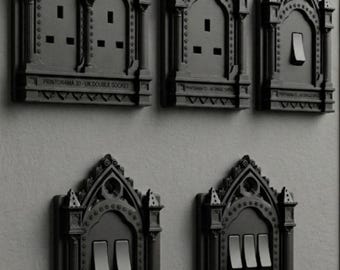 May include: A collection of five matte black electrical wall plates featuring intricate gothic architectural detailing, including pointed arches and columns. The plates include various configurations of power outlets and light switches with text labels.