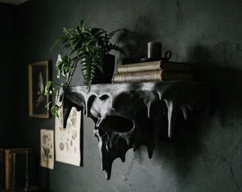 May include: A matte black wall-mounted shelf shaped like a human skull with a dripping, melted wax effect. The top surface holds a potted green fern, two stacked vintage books with worn covers, and a small dark candle holder.