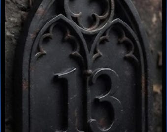 May include: A dark, weathered metal house number plaque featuring the number 13 in a raised font. The plaque has an arched top with decorative quatrefoil cutouts above and below the digits, resembling a pointed window frame design.