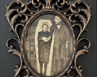 May include: An oval-shaped, ornate bronze-colored frame with intricate scrollwork detailing. Inside the frame is a vintage black and white photograph depicting a person standing next to another person lying inside a coffin-shaped structure.