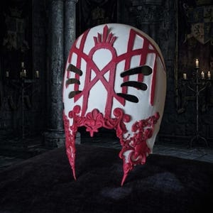 May include: A white mask featuring intricate red raised filigree and geometric patterns. The mask has two horizontal black eye slits and a prominent red crown-like emblem at the top center, with ornate red scrollwork detailing along the lower edges.