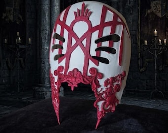 May include: A white mask featuring intricate red raised filigree and geometric patterns. The mask has two horizontal black eye slits and a prominent red crown-like emblem at the top center, with ornate red scrollwork detailing along the lower edges.