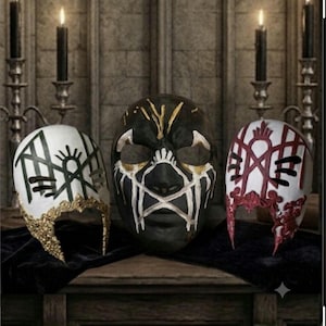 May include: Three decorative masks displayed together. The left mask is white with green geometric lines and gold filigree. The center mask is black with white and gold painted accents. The right mask is white with red lines and red filigree.