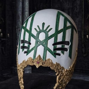 May include: A white helmet featuring a green geometric pattern and black horizontal slits. The base is adorned with intricate gold-colored filigree detailing and a small, circular red gemstone centered at the front edge of the decorative trim.