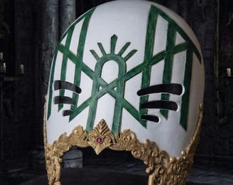 May include: A white helmet featuring a green geometric pattern and black horizontal slits. The base is adorned with intricate gold-colored filigree detailing and a small, circular red gemstone centered at the front edge of the decorative trim.