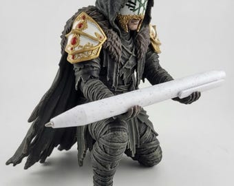 May include: A kneeling action figure wearing a dark hooded cloak, textured wrappings, and white shoulder armor with red accents. The figure wears a white mask with green markings and holds a large, white, speckled ballpoint pen.