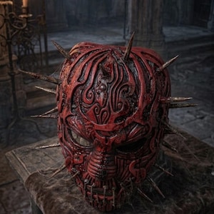 Può includere: Una maschera integrale rossa caratterizzata da intricati motivi a vortice in rilievo e numerose punte affilate dall'aspetto metallico che sporgono dai lati e dalla parte superiore. La maschera presenta una texture usurata con aree scure.