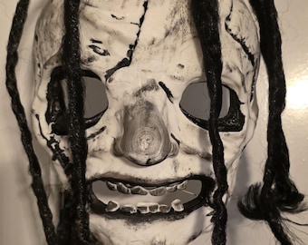 May include: A white full-face mask with a cracked, weathered texture and black detailing around the eyes and mouth. The mask features prominent black yarn dreadlocks attached to the top and sides, with visible teeth and a wide, open mouth.