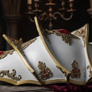 May include: A white shoulder armor piece featuring gold filigree trim and embedded red oval gemstones. The armor has a segmented, layered design with sharp, pointed edges and ornate metallic detailing along the borders and center sections.