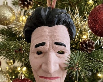 May include: A 3D-printed hanging ornament shaped like a human face with pale skin, black hair, dark eyebrows, and red lips. The surface features visible horizontal layer lines from the printing process. A thin ribbon is attached to the top for hanging.