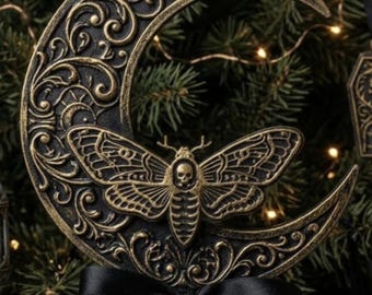 May include: A black and gold crescent moon ornament featuring an intricate moth with a skull marking on its thorax. The moon is decorated with embossed scrollwork and lunar phases, finished with a black satin ribbon tied at the base.