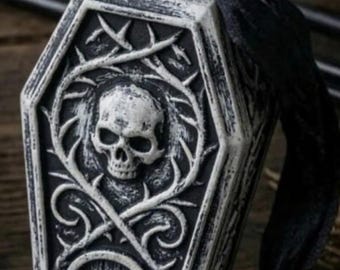 May include: A coffin-shaped ornament featuring a raised white skull and thorny vine relief on a black background. The piece includes a small arched door detail at the base and a circular loop at the top, threaded with a black ribbon for hanging.