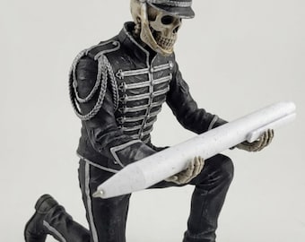 May include: A skeleton figurine dressed in a black military-style marching band uniform with silver braided accents and a tall hat, kneeling while holding a large, white, speckled ballpoint pen horizontally in its skeletal hands.