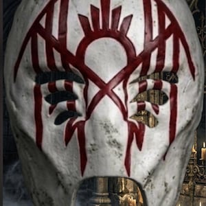 May include: A white, textured mask featuring a weathered, stone-like finish. The surface is adorned with a bold, symmetrical red graphic design that resembles stylized wings and a central crest, extending downward from the eye openings.