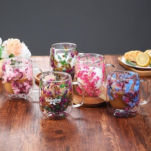 May include: A collection of clear glass mugs with double-walled construction, each filled with colorful dried flowers. One mug has the word "Valentine" printed on it. The mugs are arranged on a wooden surface.
