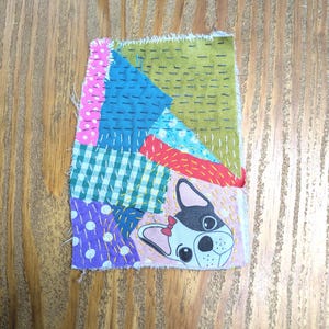 Puede incluir: Un textil de patchwork con una ilustración de bulldog francés. El diseño incorpora varios patrones de tela, incluyendo lunares, cuadros y colores lisos, cosidos con hilo visible. El bulldog tiene un lazo rojo.