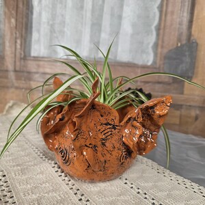 May include: A handcrafted, orange ceramic planter with a textured surface and unique wing-like details. The planter holds a green and white spider plant, adding a touch of nature to any space. The planter is sitting on a white lace tablecloth.