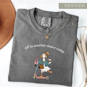 May include: Pepper-colored Comfort Colors t-shirt with the phrase "off to another dance comp." The graphic features a cartoon goose on a scooter, holding ballet shoes and a coffee cup. A gold necklace is draped across the shirt.