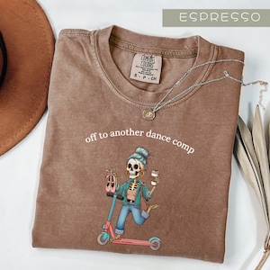 May include: Espresso-colored Comfort Colors t-shirt with the phrase "off to another dance comp" and a graphic of a skeleton riding a scooter, holding ballet shoes and a coffee cup. A silver necklace is draped across the shirt.