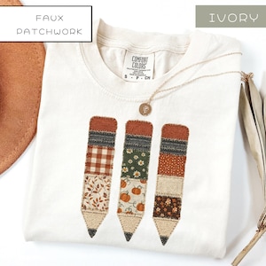 May include: An ivory t-shirt featuring a faux patchwork design of three pencils. Each pencil is composed of various fabric patterns including gingham, floral, and pumpkin prints in shades of brown, green, and cream. The shirt has a crew neck.