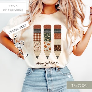 May include: An ivory t-shirt featuring a graphic of three pencils with faux patchwork patterns in autumn colors, including plaid, floral, and pumpkin prints. The text "mrs. johnson" is printed in brown script below the pencil graphic.