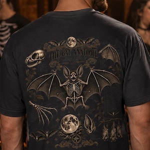 May include: A dark grey short-sleeve t-shirt featuring a detailed graphic on the back. The design displays a central bat skeleton with outstretched wings, surrounded by smaller illustrations of a skull, a moon, and hanging bats with the text The Bat Anatomy.