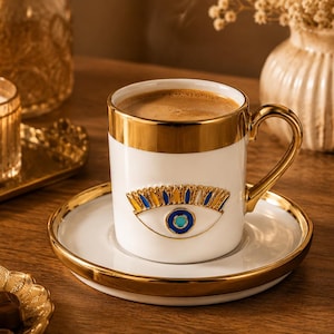 Könnte beinhalten: Weiße Kaffeetasse und Untertasse mit goldenem Rand und Augendesign. Die Tasse ist mit Kaffee gefüllt und steht auf einer passenden Untertasse. Das Set steht auf einer Holzoberfläche.