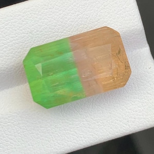 May include: A rectangular faceted gemstone featuring a distinct bicolor transition from a vibrant lime green on the left side to a soft peach or light orange hue on the right side, showcasing internal clarity and a polished, reflective surface.