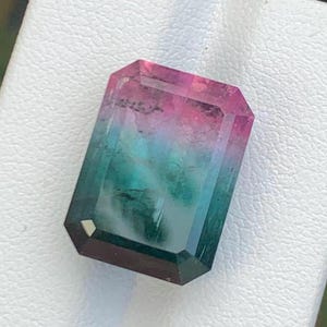 May include: A rectangular faceted gemstone featuring a gradient colour transition from a vibrant pink at the top to a deep teal and green at the bottom. The stone has a polished surface with visible internal inclusions and sharp, clean edges.