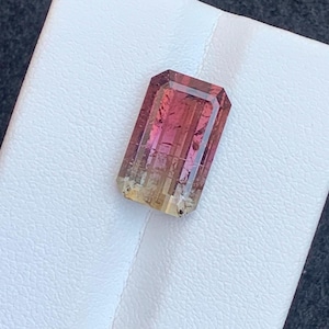 May include: A rectangular gemstone with a gradient of colours, transitioning from a yellow-orange hue at the base to a vibrant pink at the top. The gem has a faceted cut, showcasing its internal structure and clarity. The stone is set against a white surface.