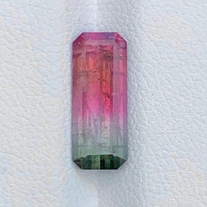 May include: A rectangular, emerald-cut gemstone featuring a distinct colour gradient that transitions from a deep, vibrant pink at the top to a soft, translucent green at the base, showcasing internal facets and a polished, reflective surface.