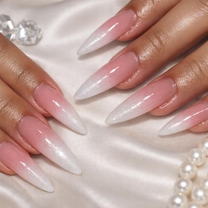 May include: Close-up of a set of long, pointed acrylic nails with a pink and white ombre design. The nails have a glossy finish and are set against a white satin background. A few decorative elements are visible.