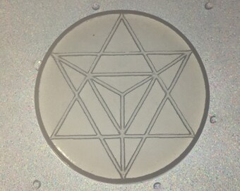 May include: A circular, off-white object with a geometric design of interconnected triangles and a star shape. The design is etched in a gray color. The object is set against a shimmering, light-colored background.